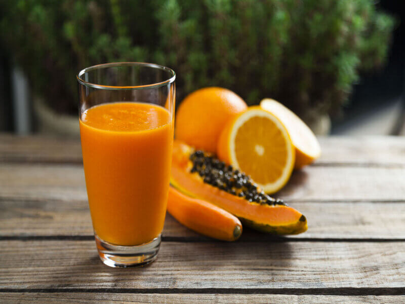 Copo de vidro com suco laranja em cima de uma mesa com pedaço de mamão, laranja e uma cenoura