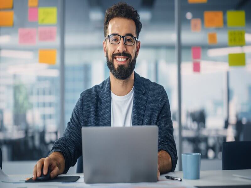 Homem de óculos, sentado sorrindo e mexendo em um notebook