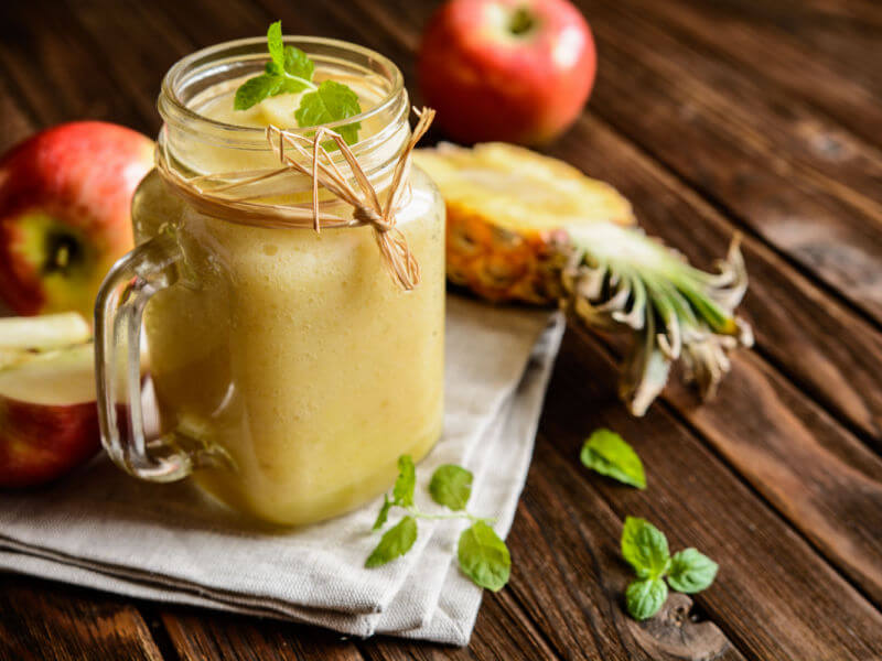 Vitamina amarela em copo de vidro com maçã e abacaxi ao lado