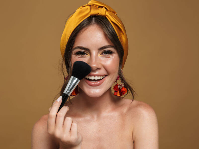Mulher de tiara amarela no cabelo sorrindo e segurando pincel de maquiagem no rosto