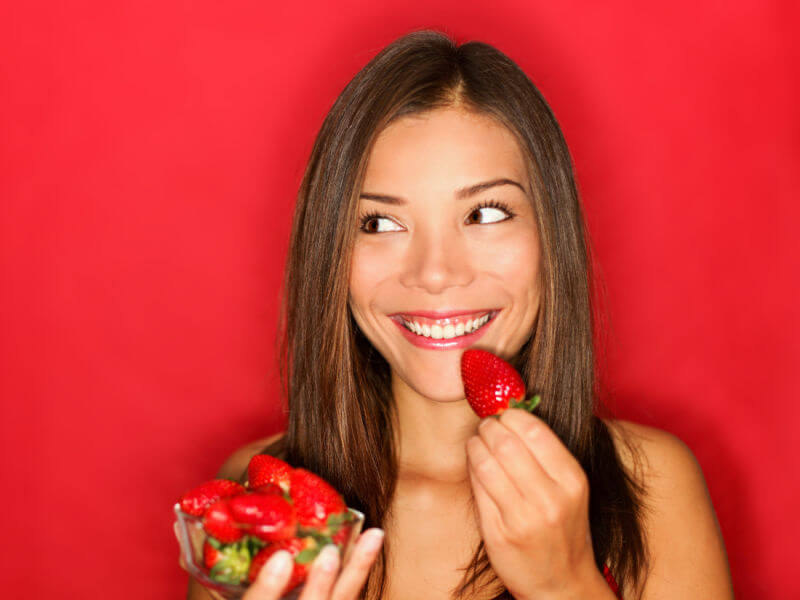 Mulher sorrindo segurando um recipiente com morangos em frente a um fundo vermelho