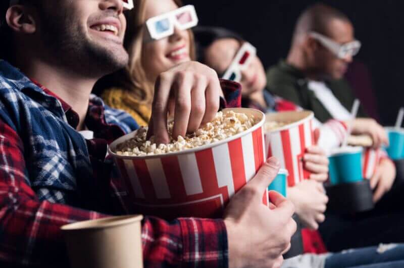 Jovens amigos sorridentes em óculos 3d comendo pipoca e assistindo filme no cinema