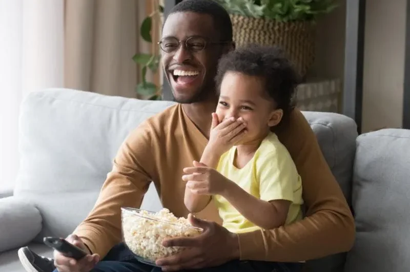 Pai e filho assistem a televisão sorridentes, e com pipoca.