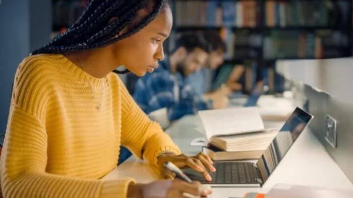 Na imagem, jovem estudante em uma biblioteca escrevendo no computador e em um caderno concentrada