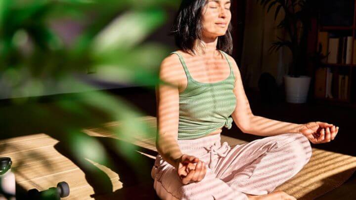 Mulher de regata verde e calça rosa meditando ao lado de uma planta