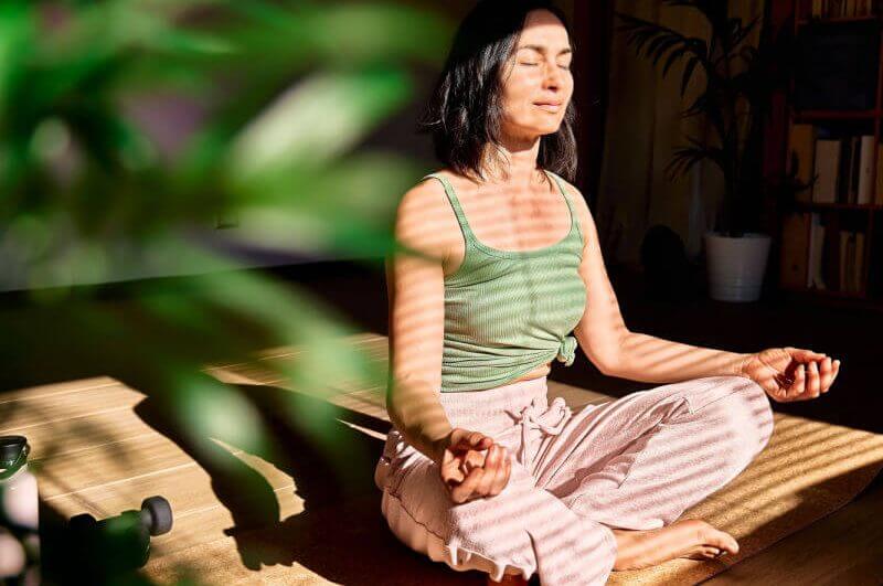 Mulher de regata verde e calça rosa meditando ao lado de uma planta