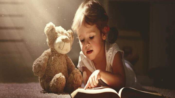 Menina deitada lendo livro para um urso de pelúcia