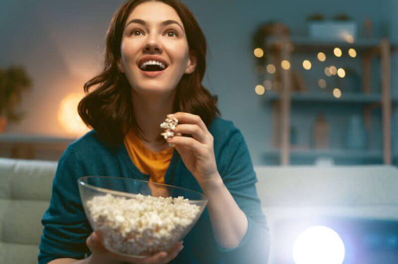 Jovem mulher assistindo projetor, TV, filmes com pipoca à noite. Garota passando um tempo em casa.