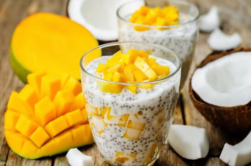 Copo de vidro com pudim de chia e pedaços de manga em cima de uma mesa de madeira