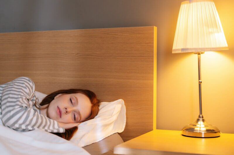 Na imagem, mulher dorme em uma cama com cabeceira de madeira. Ao lado, uma mesa de canto e um abajur com lâmpada de tom quente e amarela