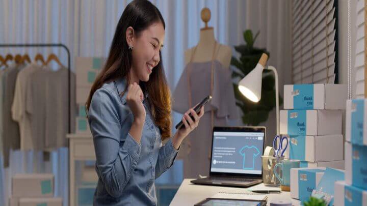 Mulher sentada mexendo no celular com computadores e caixas ao seu redor