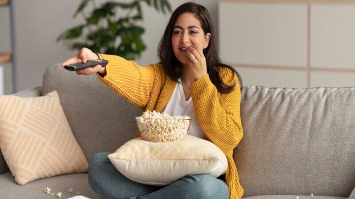Mulher de casaco amarelo sentada em um sofá bege comendo pipoca