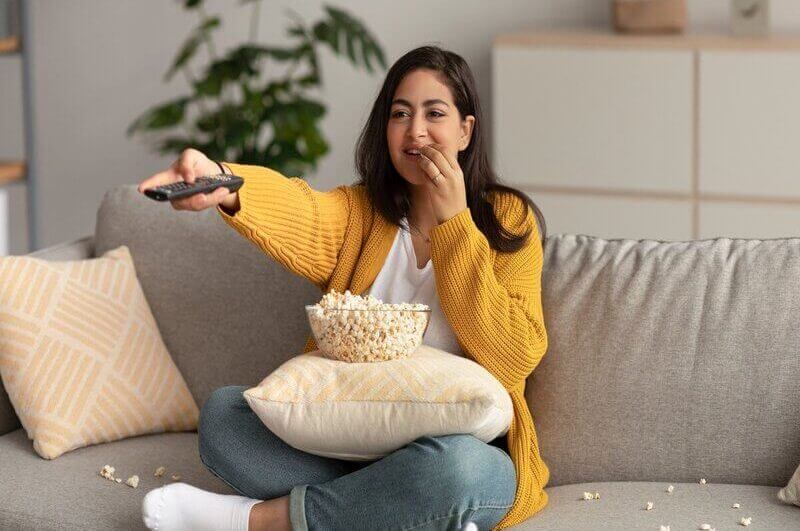 Mulher de casaco amarelo sentada em um sofá bege comendo pipoca