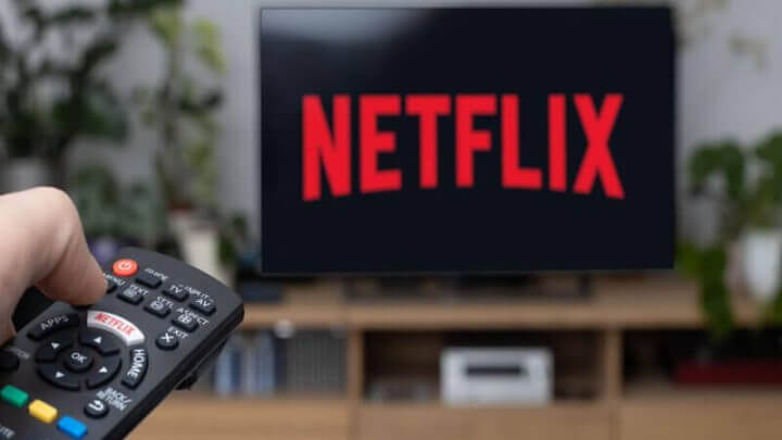 TV com logo da Netflix em rack marrom. Mão com controle remoto apontado para a TV