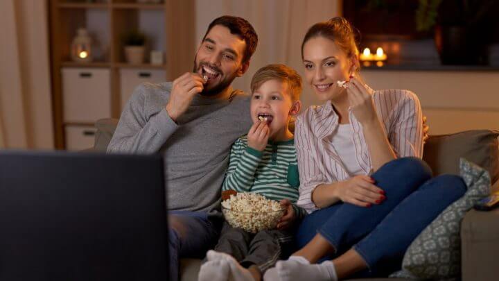 Pai, filho e mãe sentados no sofá vendo filme e comendo pipoca