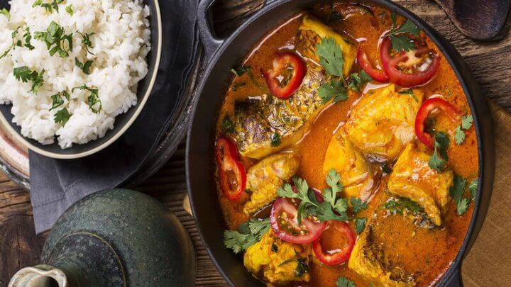 Mesa com uma travessa de cerâmica arroz branco, uma panela grande de ferro com moqueca de peixe e um jarro de cerâmica cinza