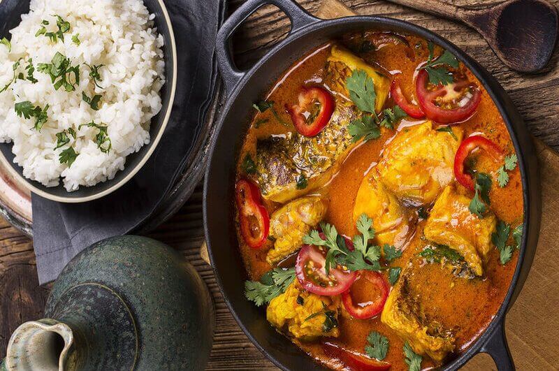 Mesa com uma travessa de cerâmica arroz branco, uma panela grande de ferro com moqueca de peixe e um jarro de cerâmica cinza