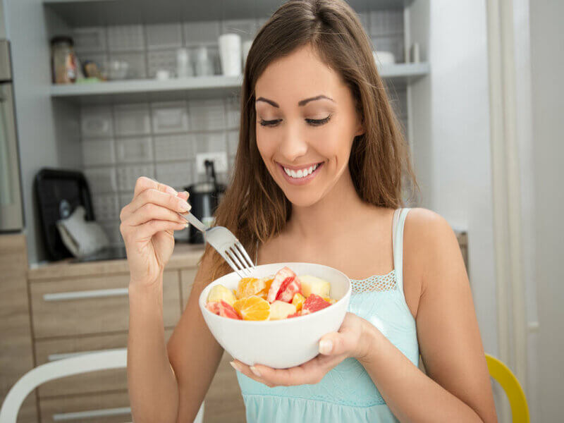 Mulher de cabelos longos sorrindo e segurando um recipiente branco com frutas