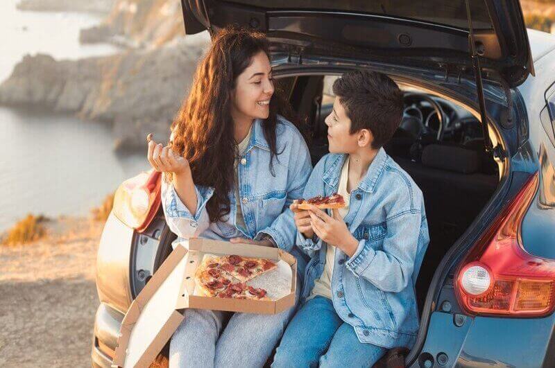 Mulher e menino sentados no porta malas de um carro comendo pizza