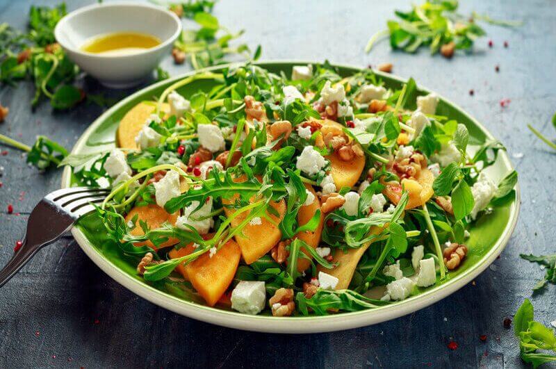 Salada com folhas de rúcula, caqui, queijo feta e nozes em um prato grande