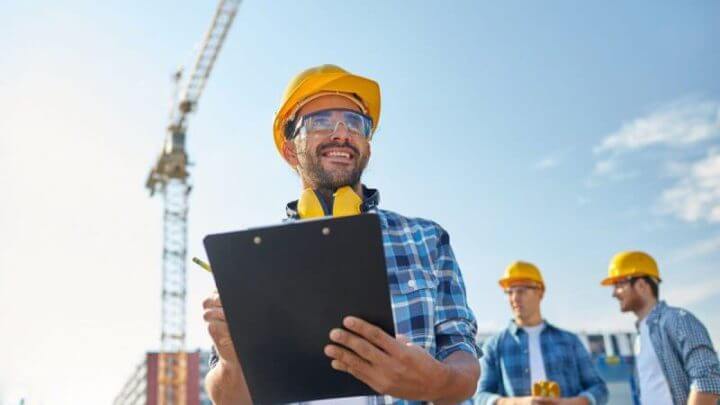 Chefe de construção com prancheta na mão; logo atrás tem mais outros dois trabalhadores em pé conversando; na imagem, todos estão de blusa xadrez azul e com capacete amarelo