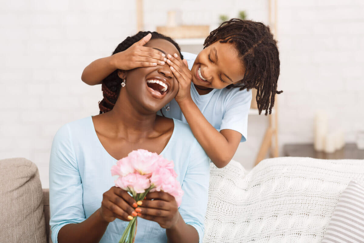 Filha tapando os olhos da mãe, que segura um buquê de flor