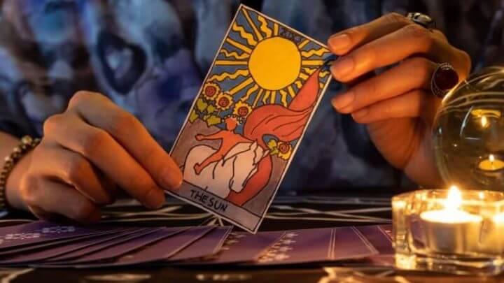 Cartomante de mãos segurando o cartão THE SUN e cartas de tarô na mesa perto de velas acesas à luz de velas