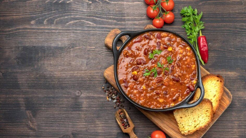 Chilli vegano servido em recipiente rústico preto em cima de tábua de madeira com pães, pimenta e especiarias em volta.