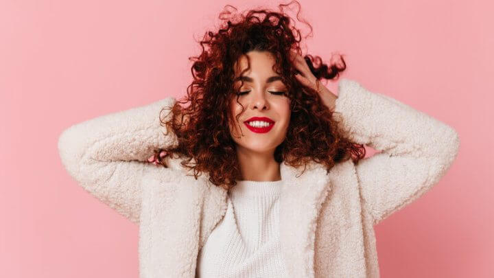 Mulher sorrindo com as mãos no cabelo em frente a um fundo rosa