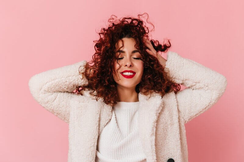 Mulher sorrindo com as mãos no cabelo em frente a um fundo rosa