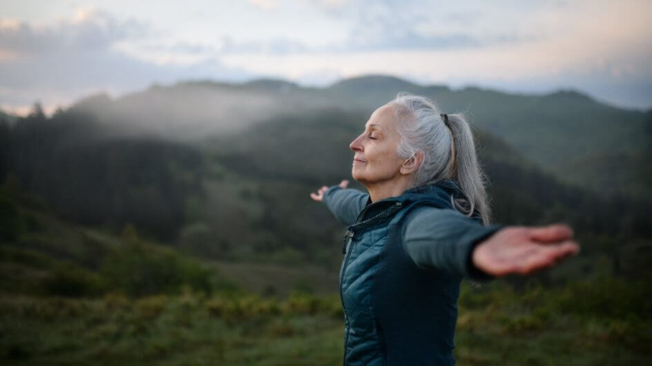 Mulher de braços abertos e olhos fechados em cima de montanha.