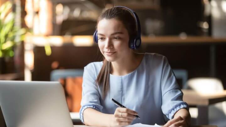 Jovem de fones de ouvido concentrada estudando em ambiente comum com computador e realizando anotações