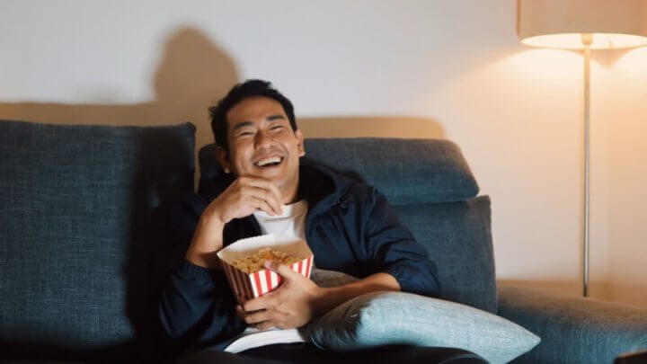 Homem alegre sentado no sofá e assistindo filme com pipoca.
