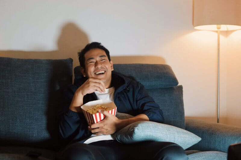 Homem alegre sentado no sofá e assistindo filme com pipoca.