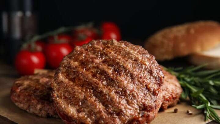Hambúrguer de carne em cima de folha marrom. Tomates e alecrim ao fundo