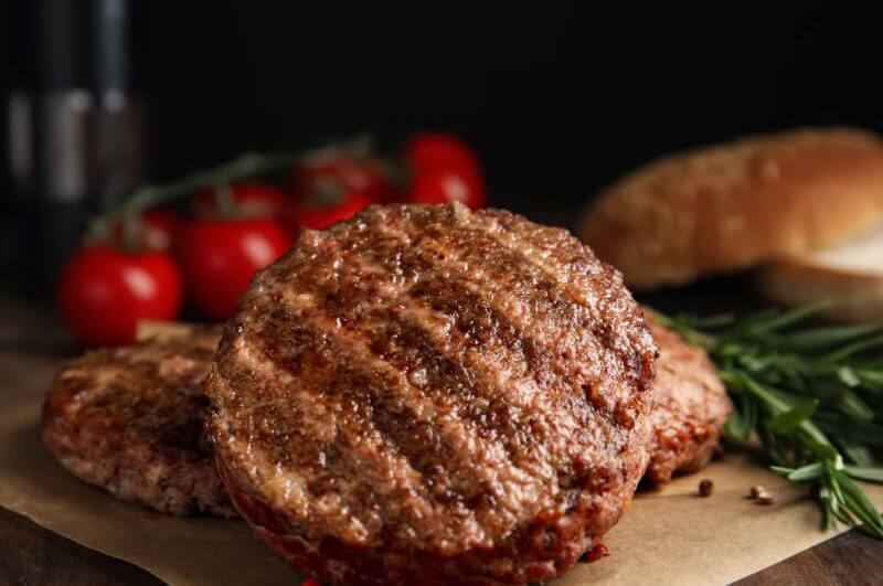 Hambúrguer de carne em cima de folha marrom. Tomates e alecrim ao fundo