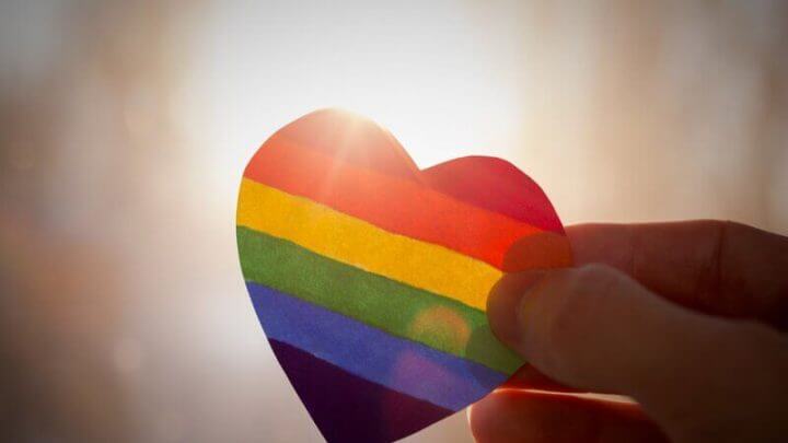 Uma mão segurando um coração colorido com as cores da bandeira LGBTQIA+