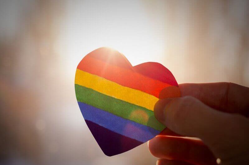 Uma mão segurando um coração colorido com as cores da bandeira LGBTQIA+