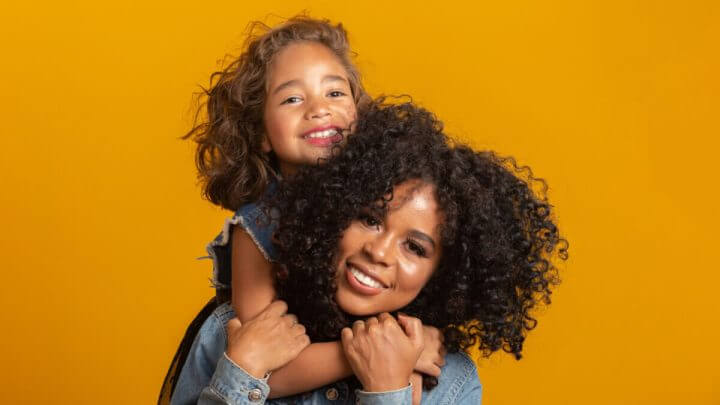 Mãe com filha apoiada nas costas em fundo amarelo
