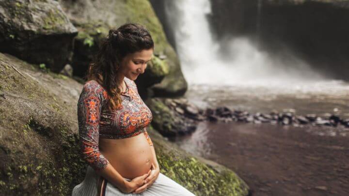 Mulher grávida em cachoeira com as mãos na barriga e olhando para baixo, sorrindo.