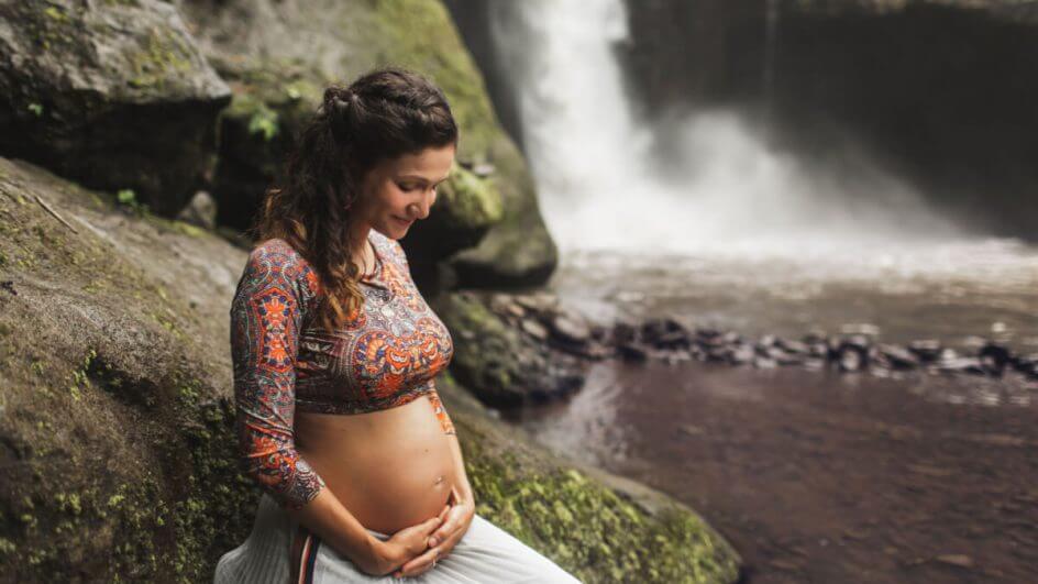 Mulher grávida em cachoeira com as mãos na barriga e olhando para baixo, sorrindo.
