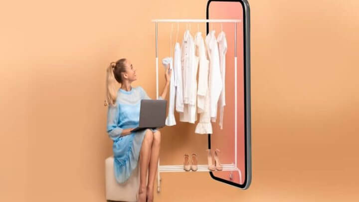 Mulher sentada com notebook no colo e roupas penduradas em cabideiro ao lado