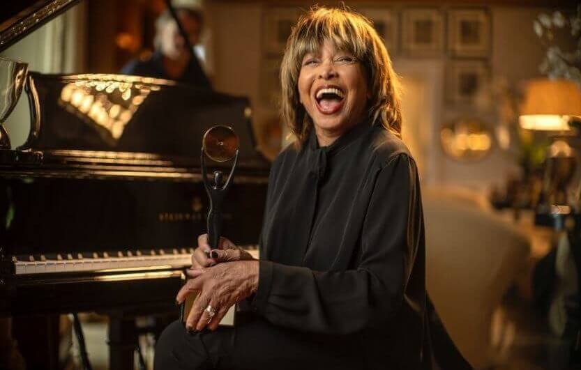 Tina Turner sorrindo ao lado de piano.