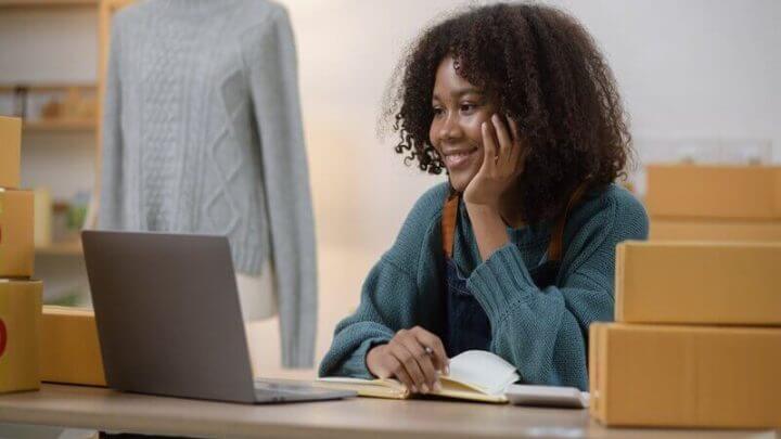 Mulher sentada sorrindo olhando para um notebook com caixas ao seu redor