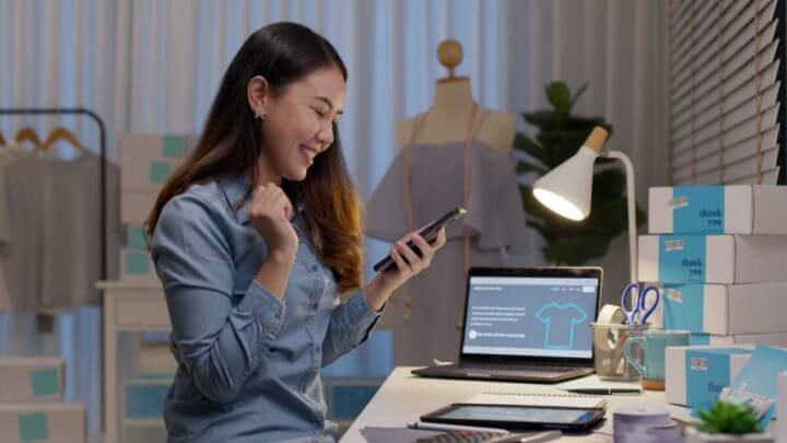 Mulher sentada mexendo no celular com computadores e caixas ao seu redor