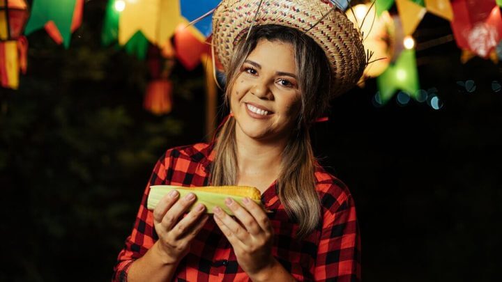 Mulher com chapéu e camisa xadrez sorrindo segurando uma espiga de milho