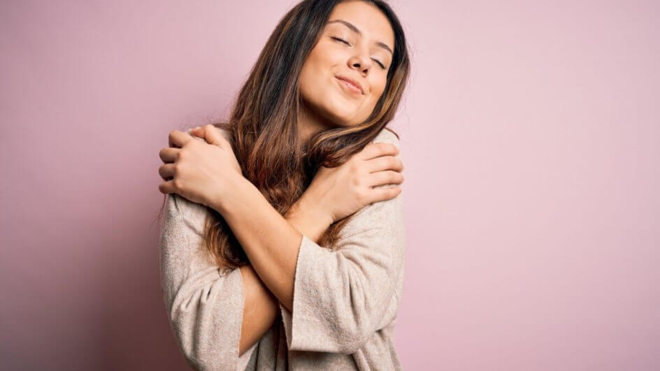 Mulher vestindo suéter casual em pé sobre fundo rosa abraçando-se feliz