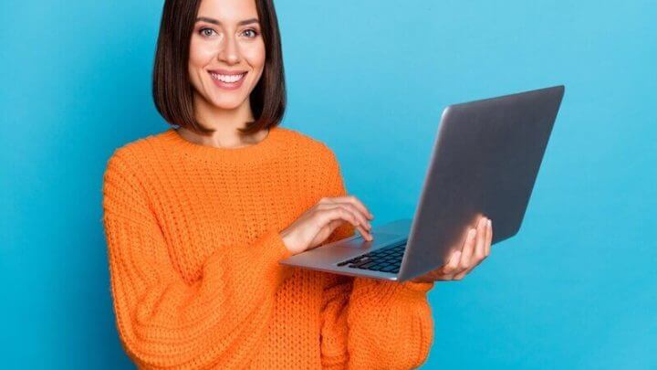 Mulher de blusa laranja sorrindo e segurando um notebook