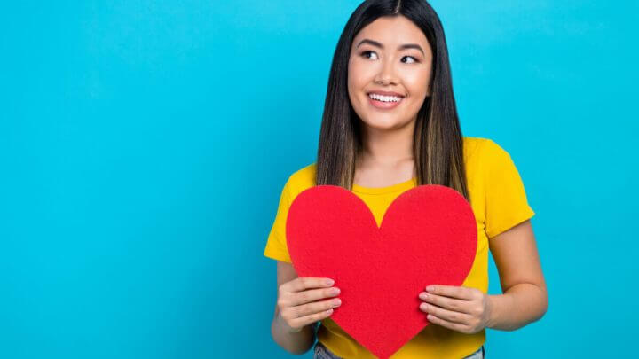 Menina sorridente de blusa amarela em fundo azul segurando coração de papel vermelho