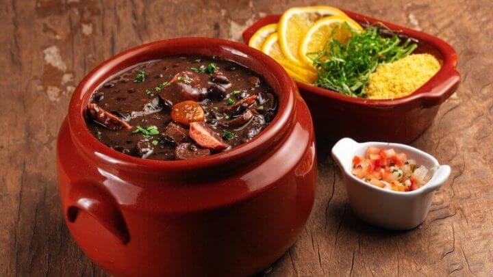 Recipiente de vidro marrom com feijoada e linguiça em cima de uma mesa de madeira com dois recipientes contendo laranja e tomate